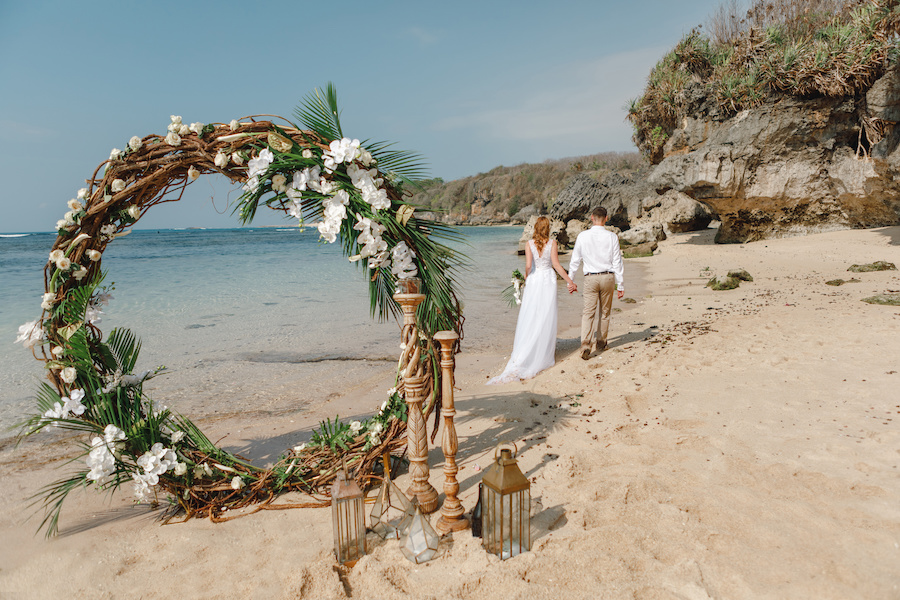 Photo of a beach wedding arch woven out of branches and decorated with palm fronds and tropical flowers