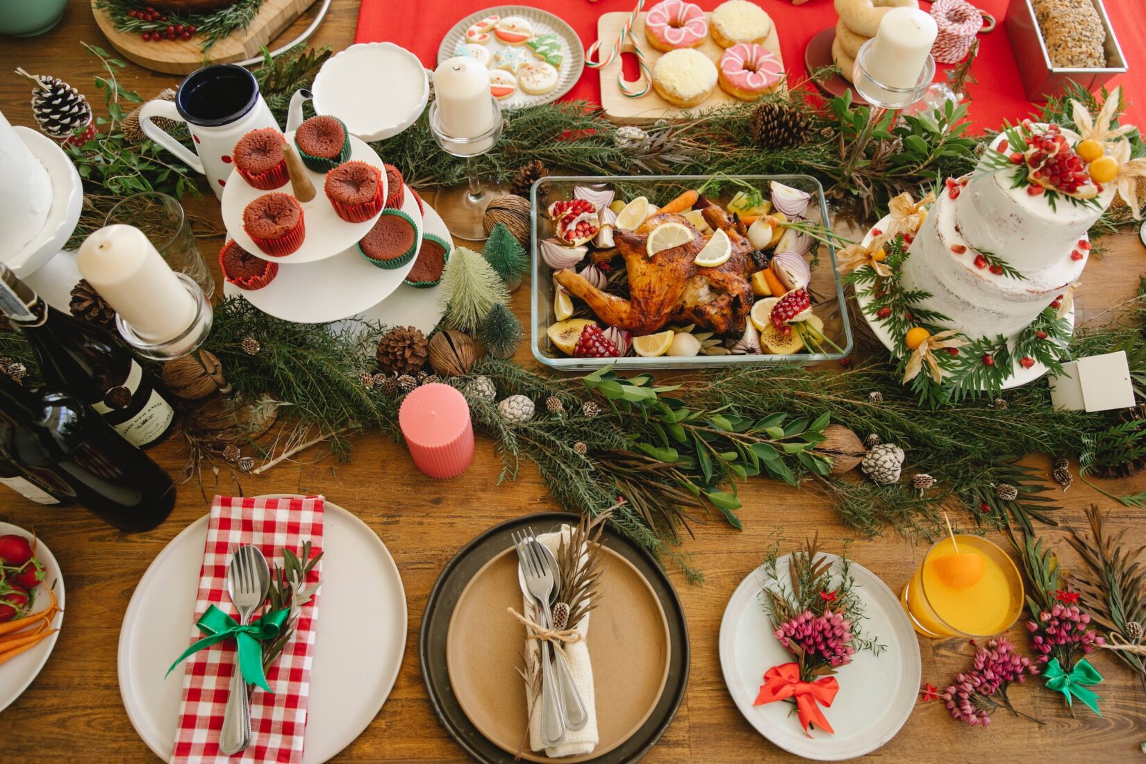 A holiday table with a roasted chicken centerpiece, surrounded by evergreen decorations, a layered cake, cupcakes, and colorful table settings.