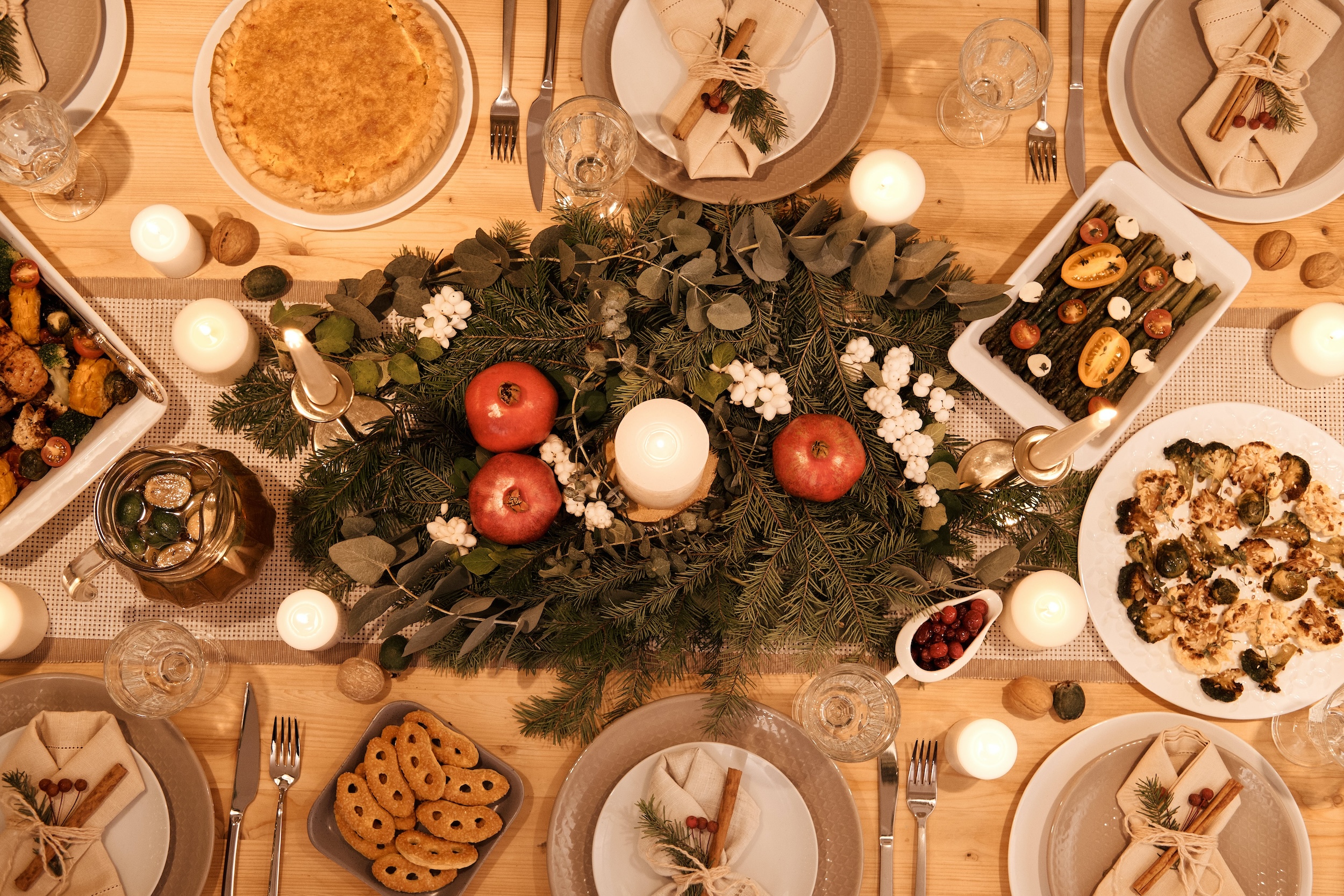 A festive table setting featuring a holiday centerpiece with candles, a pie, roasted vegetables, all under soft lighting.