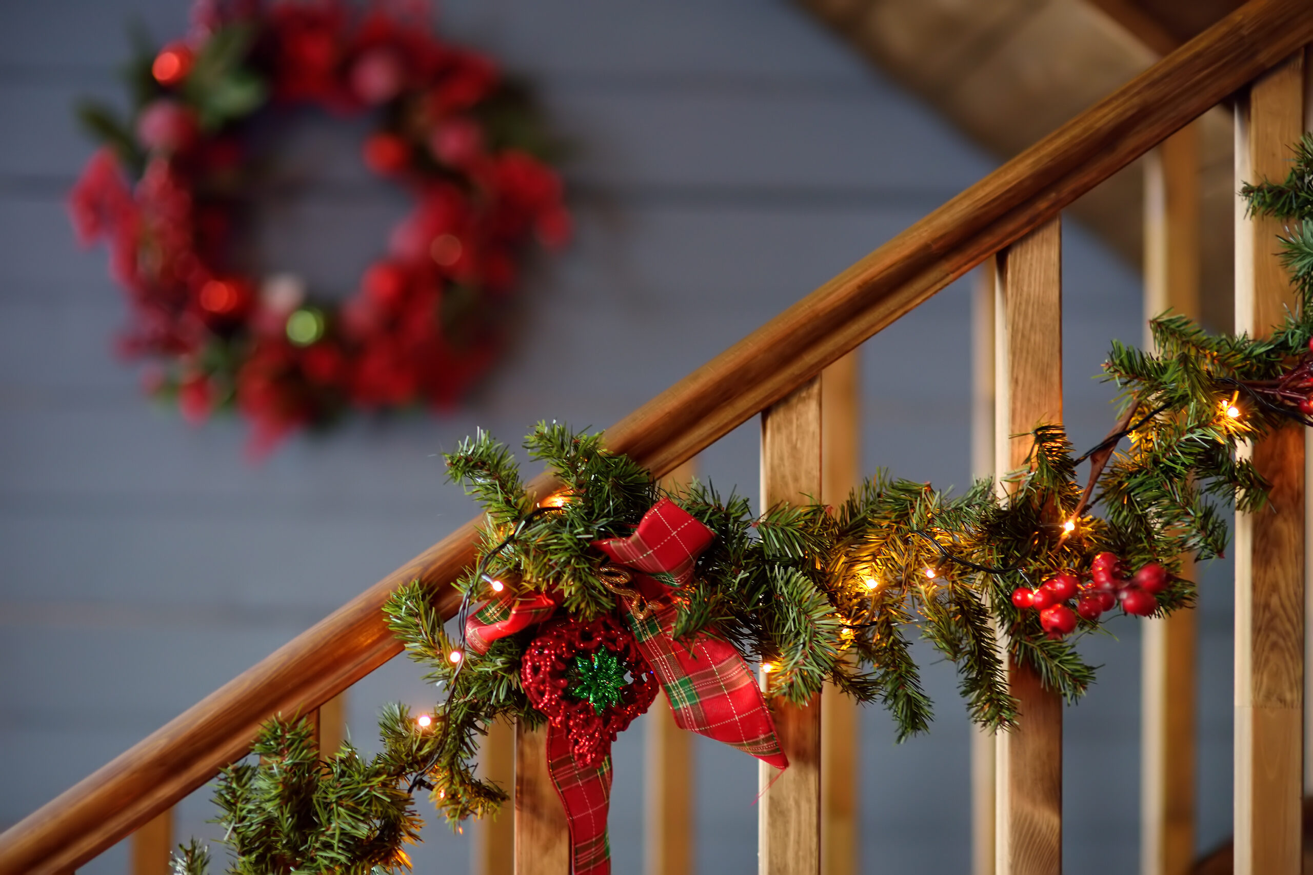 A warmly lit cedar garland adorned with red ribbons and festive ornaments winding down a wooden staircase railing.