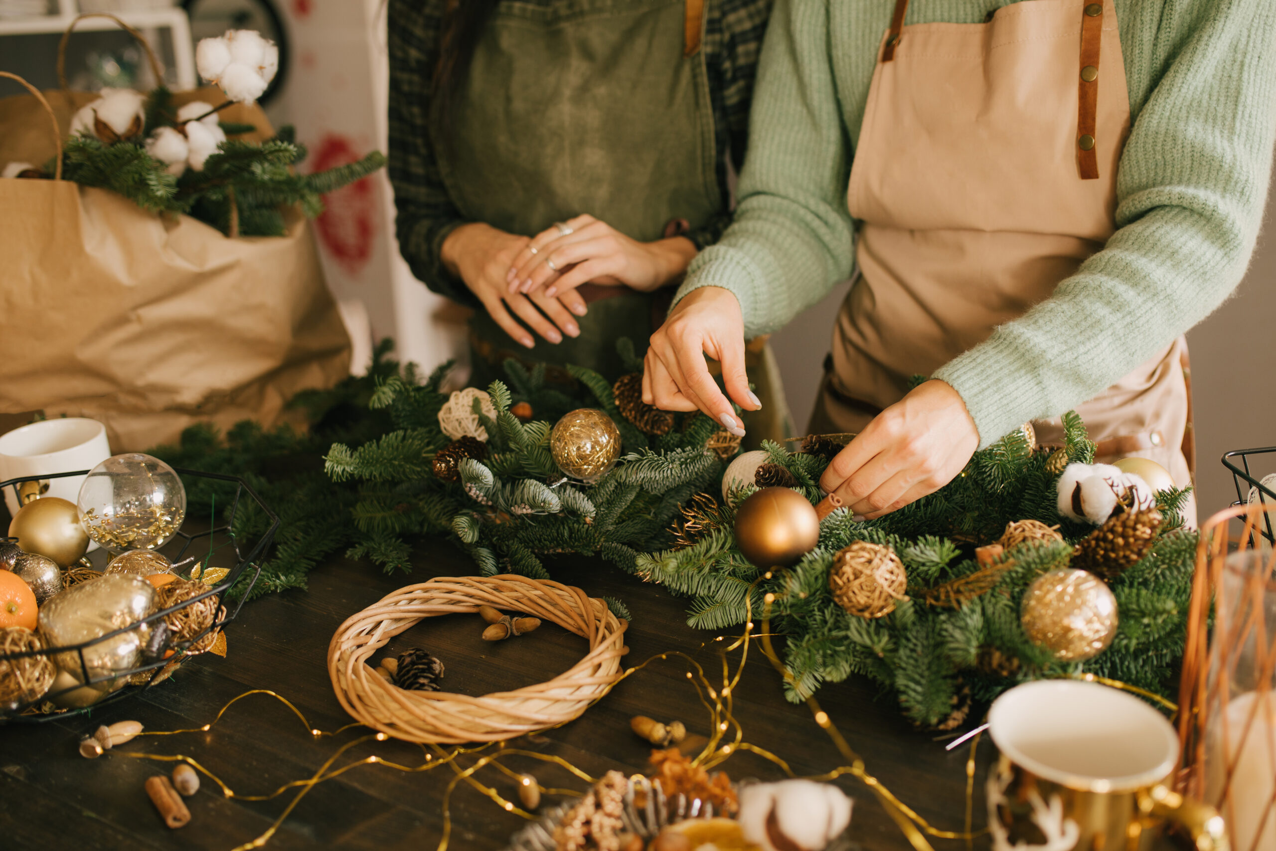 Two people in aprons adding the finishing touches to a luxurious cedar garland decorated with gold baubles, pinecones and twinkling lights.
