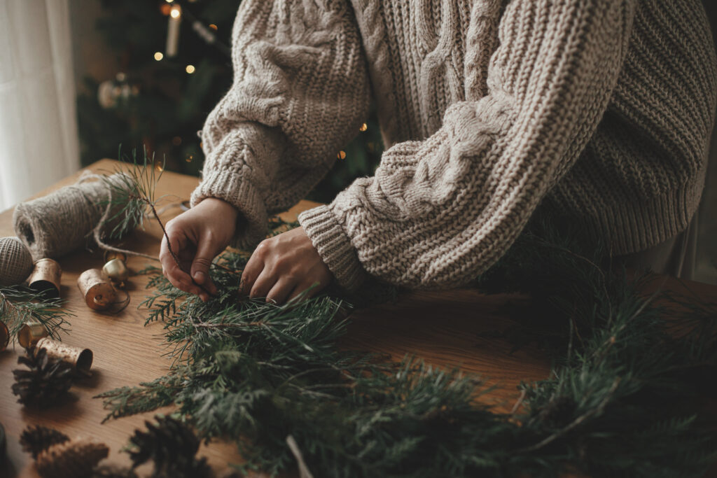 A close-up of hands arranging natural elements like pinecones and greenery into a cedar garland, set against a cozy backdrop.