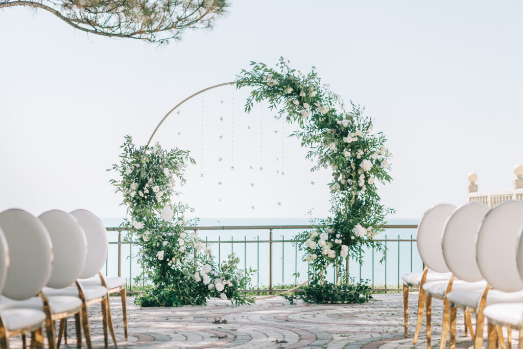 A circular wedding arch decorated with greenery and white flowers set against a seascape backdrop.