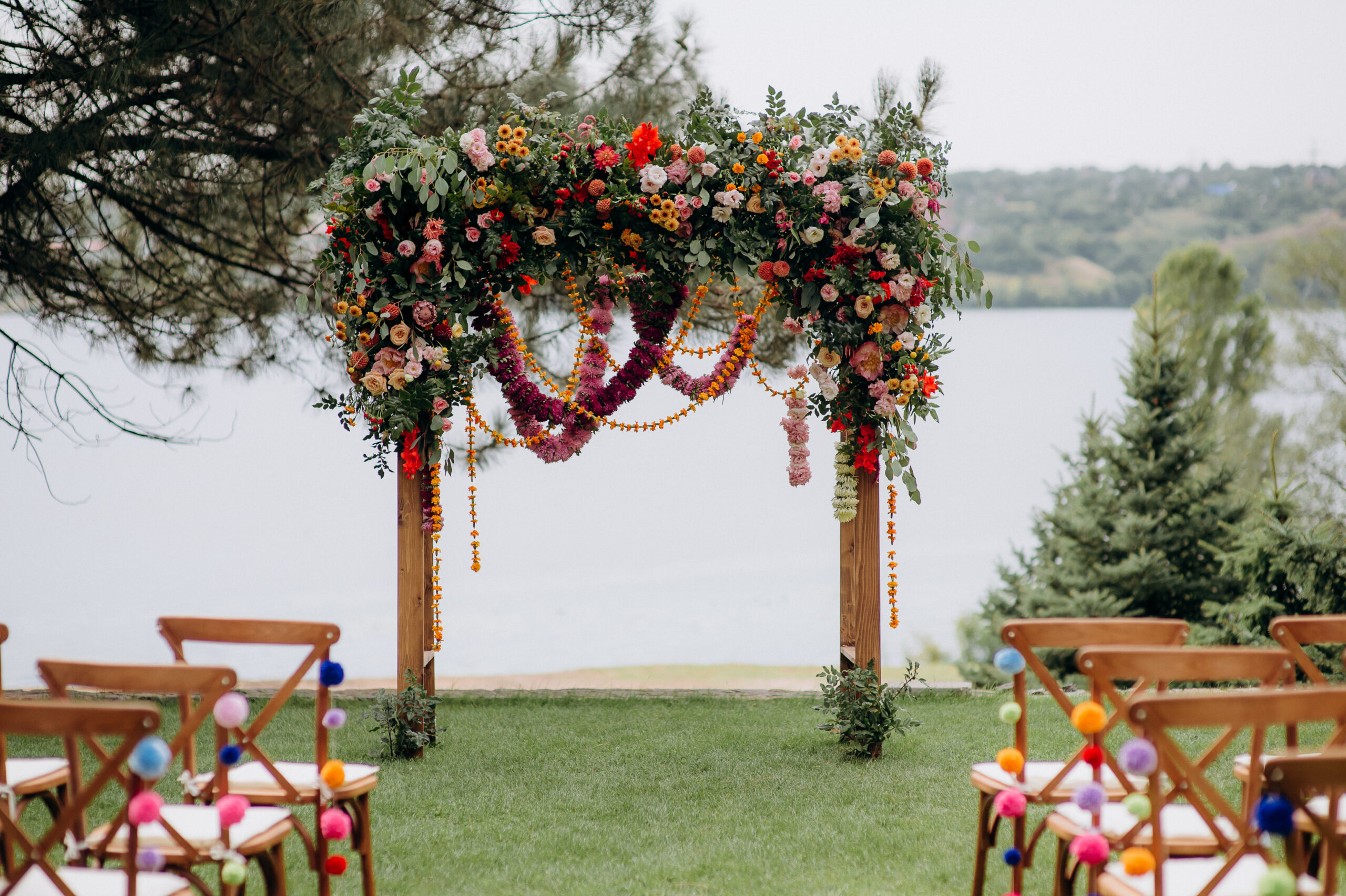 A wedding arch with lush greenery and accents of vibrant pink, orange, and yellow flowers set by a lakeside.