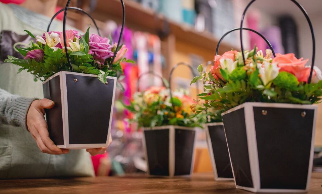 A hand holding a black square pot with a spring arrangement of pink and white flowers and green ferns, ready for gifting.