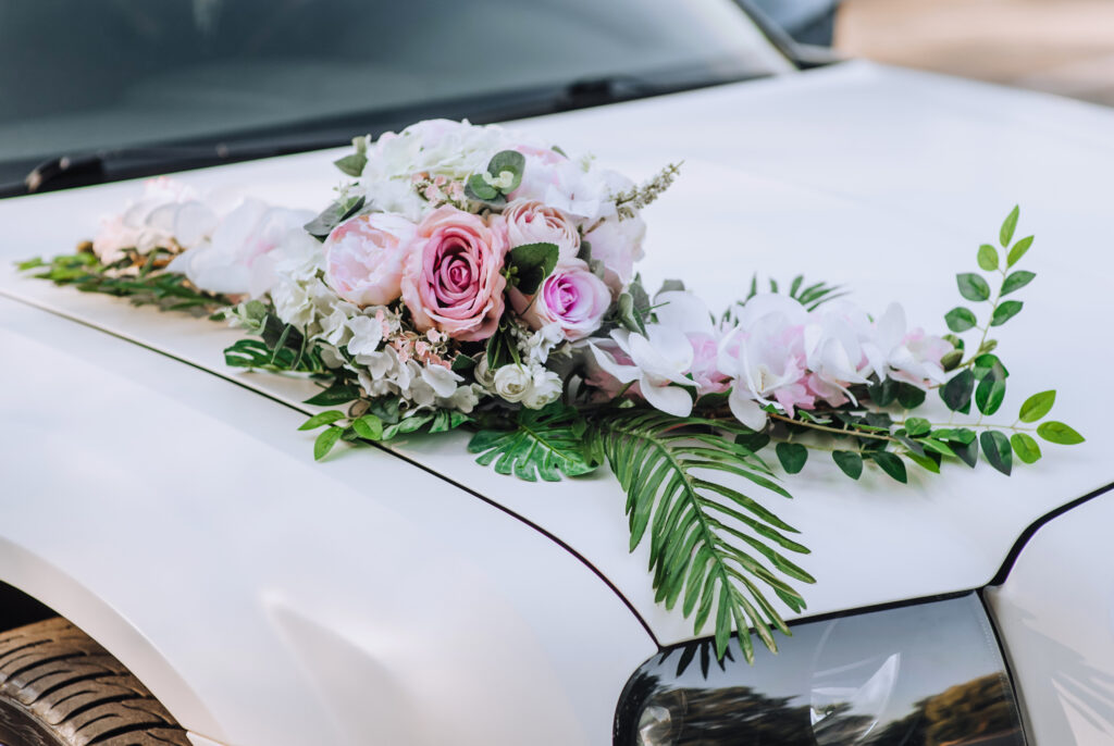 A spring floral arrangement featuring pink roses and white blossoms, accented with green fern leaves, laying on top of the hood of a white car.