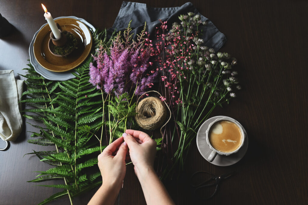 Top-down shot of a person's hands as they arrange a spring arrangement of purple and pink flowers and ferns on a dark wooden table with a lit candle and a cup of coffee nearby.